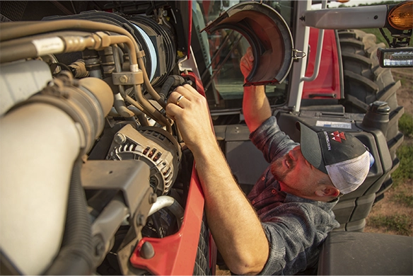 A technician wearing a cap and plaid shirt works on the engine of a red tractor, lifting the hood and inspecting components